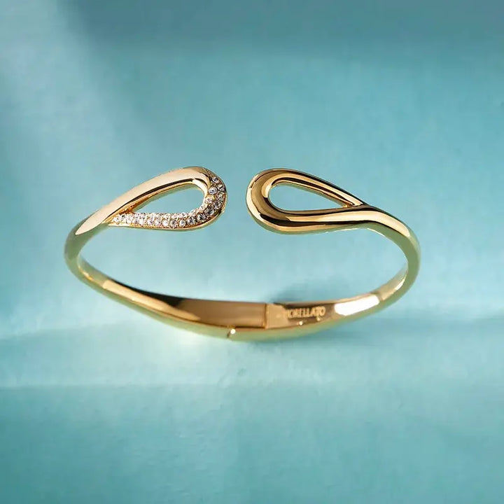 Лайфстайл кадър на позлатена гривна bangle със скулптурен отворен дизайн върху светлосин фон.