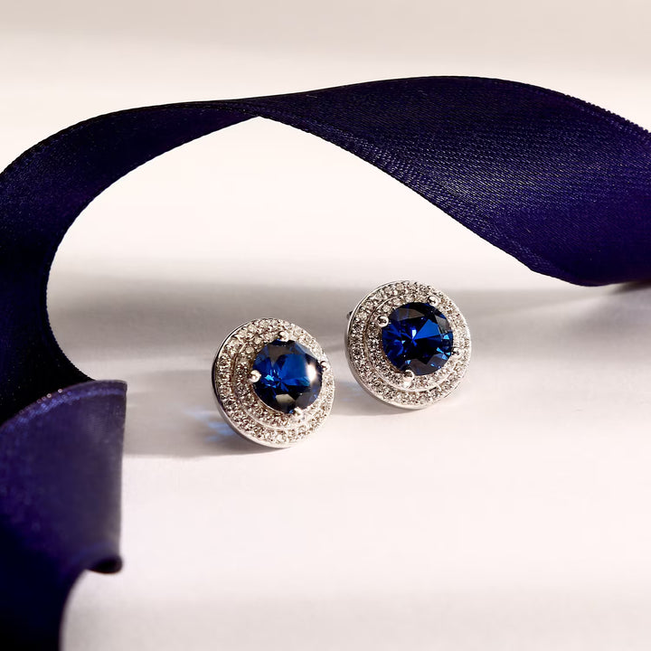 Лайфстайл кадър на сребърни обеци със син циркон върху светъл фон с лилава лента.