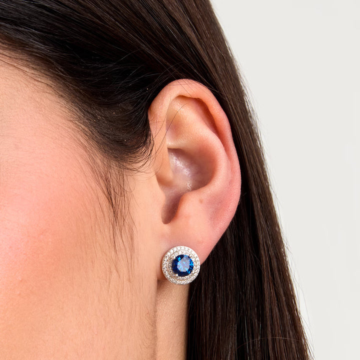 Близък план на ухо с обеца „ореол“ – син център и бели циркони.

