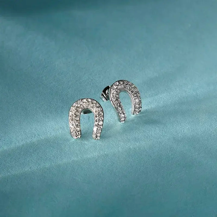 Лайфстайл кадър на сребристи обеци „подкова“ с кристали върху светлосин фон.